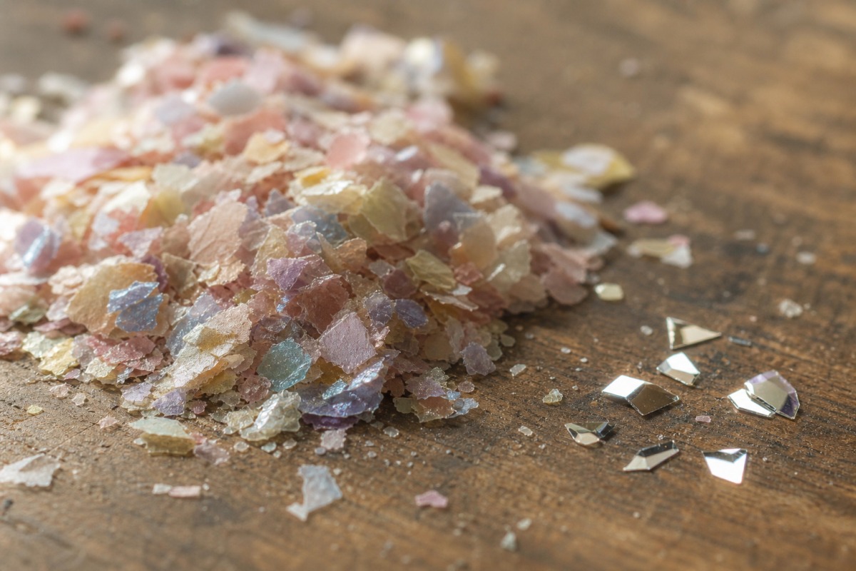 Detail van een hoopje matte cellulose-bioglitter naast enkele glanzende synthetische PET-deeltjes op een houten oppervlak.